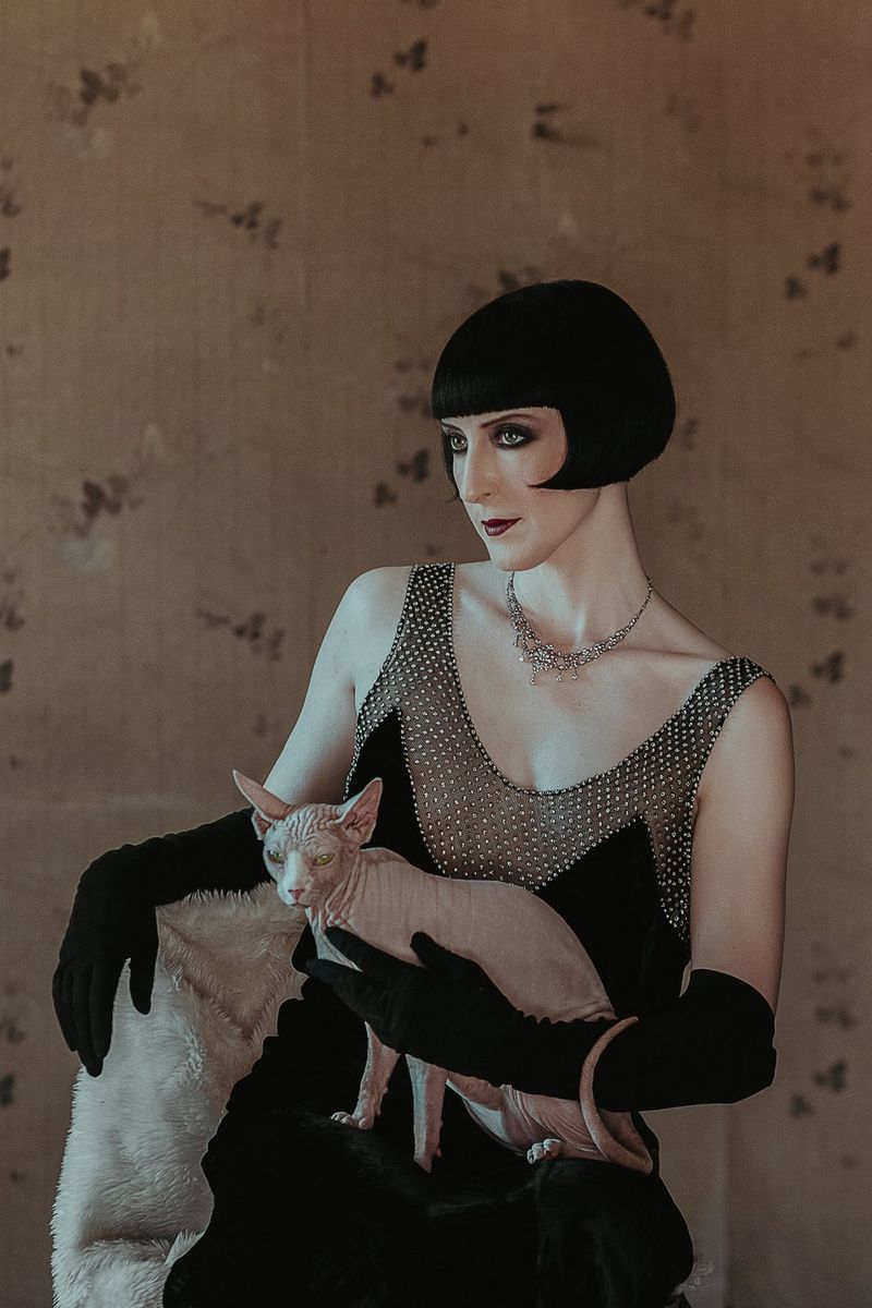 A woman with a short black bob and dressed in 1920s clothing holds a small Sphynx cat. 