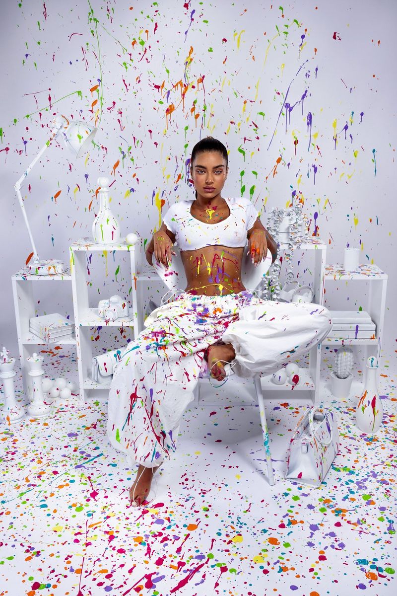 A woman in a white crop top and white baggy trousers poses on a chair in a white room splattered with paint, in a fashion portrait taken by Mor Tzidon on a Canon EOS R5.
