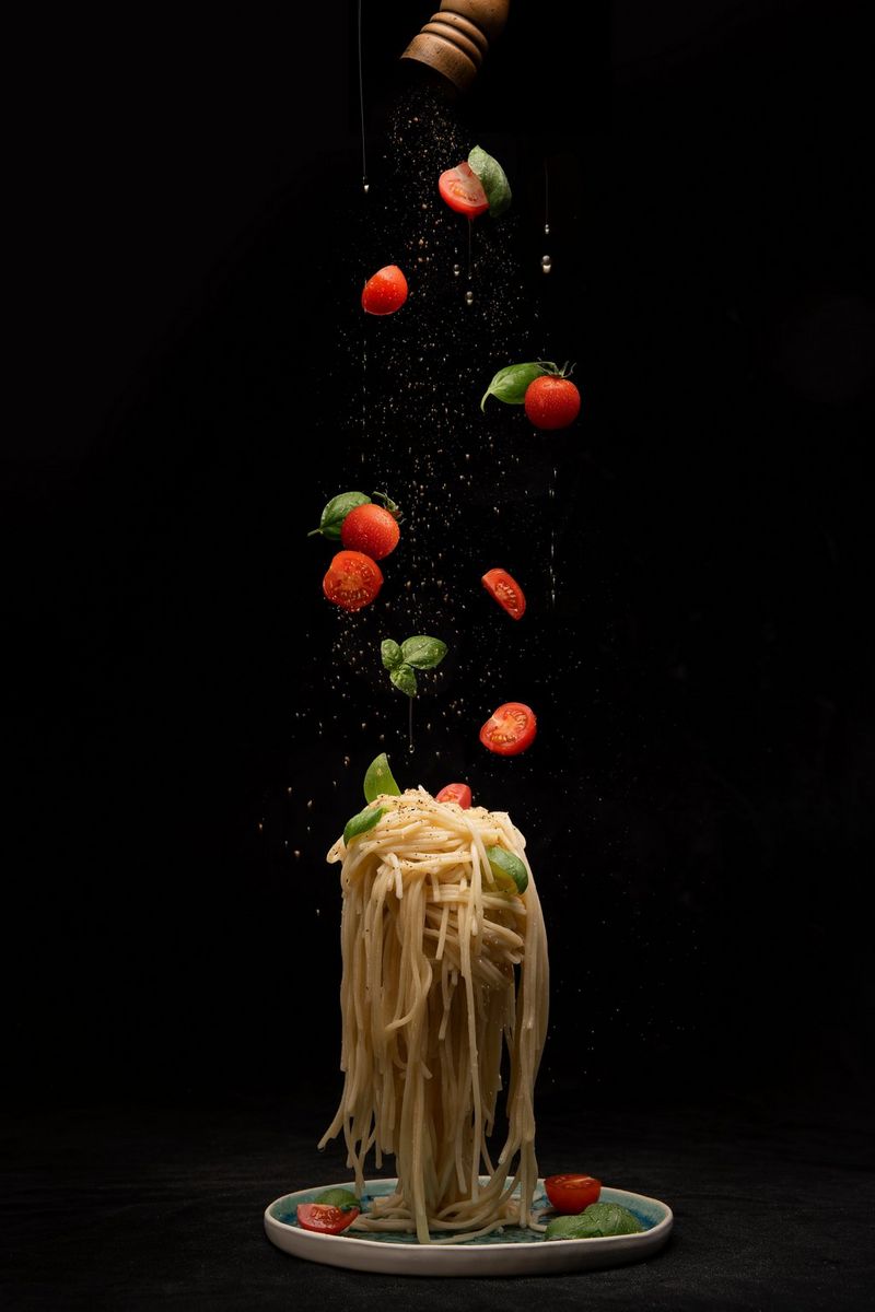 A photo of spaghetti falling down onto a plate. Ingredients above look like they're being dropped on the spaghetti. Taken against a black background on a Canon EOS R5 by Naim Chidiac.