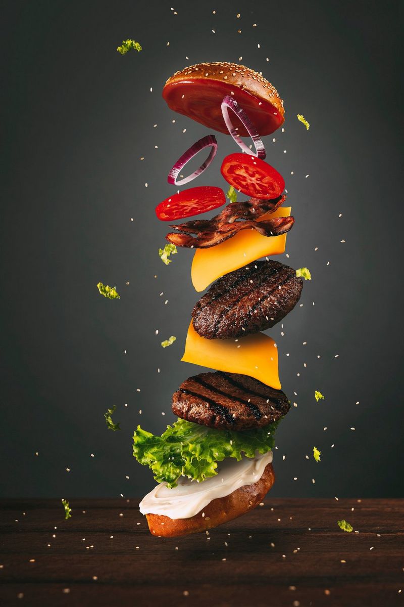 A photo of a burger in mid air, with the individual layers of vegetables and slices of cheese visible. Taken on a Canon EOS R5 by Naim Chidiac.