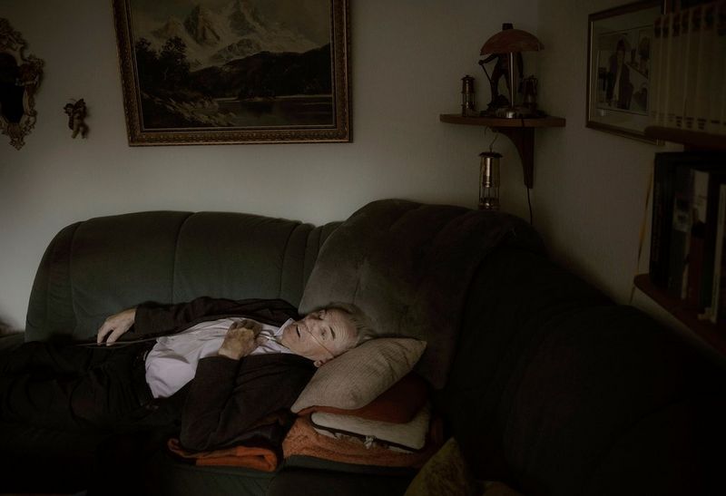 An older man wearing a breathing tube lies down on a sofa. 