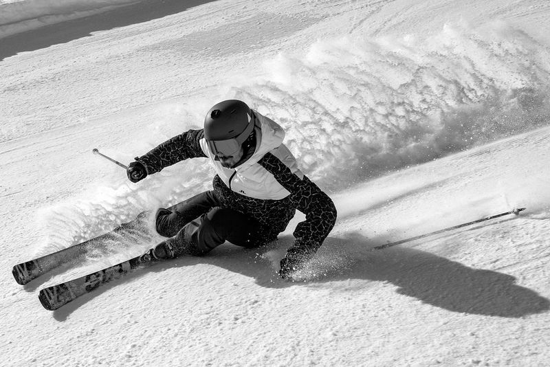 A black and white close up photograph of a male skier speeding down a mountain, leaving a trail of powdered snow in his wake. Taken on a Canon EOS R5 by Nico Schaerer.