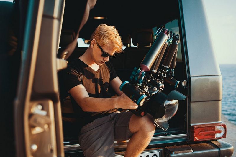 A man wearing shorts, sunglasses and a t-shirt and leaning against a car boot, adjusts his diving and camera equipment.
