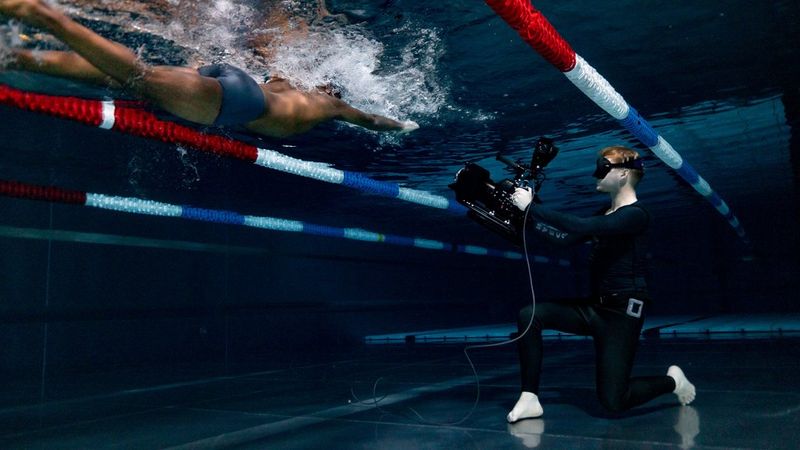 A man wearing a wetsuit and mask and kneeling on the floor of a pool, films a swimmer from below.