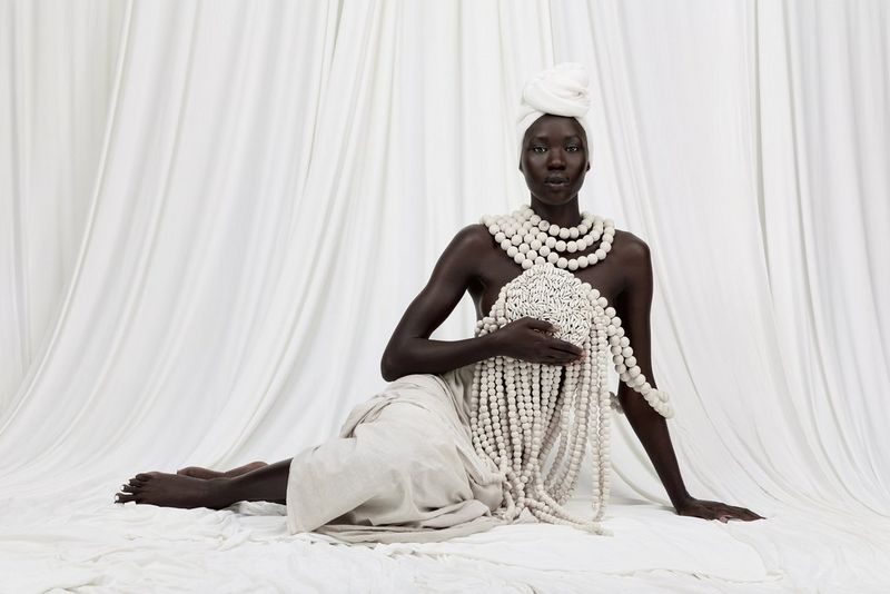 A seated model, wearing a cream turban and draped in natural linen, holding a circle of cowrie shells attached to long strings of cream beads.