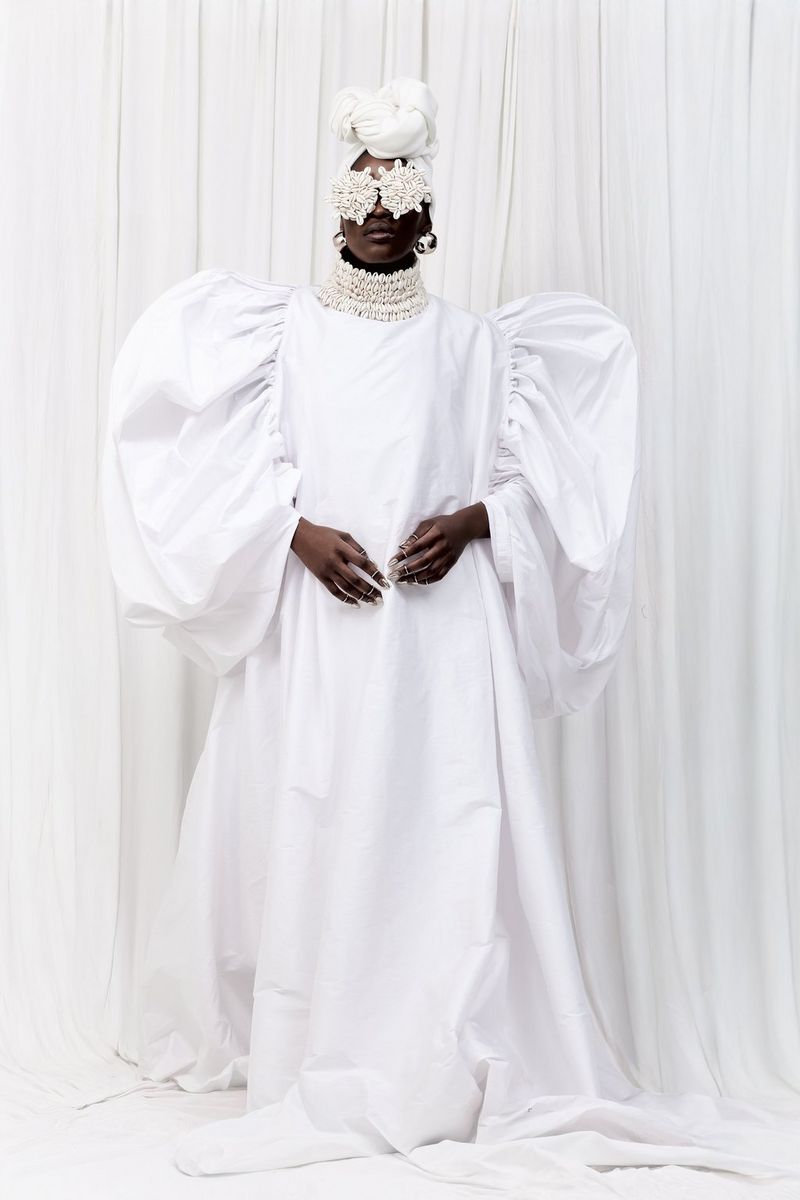 A model in a voluminous white dress wearing a cowrie shell necklace with matching eye coverings.