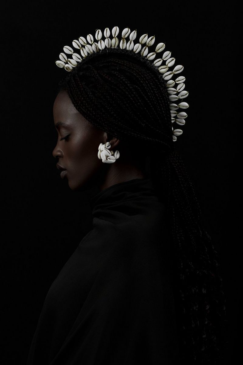 A darkly lit side profile portrait of a model wearing a cowrie shell earring and headpiece.