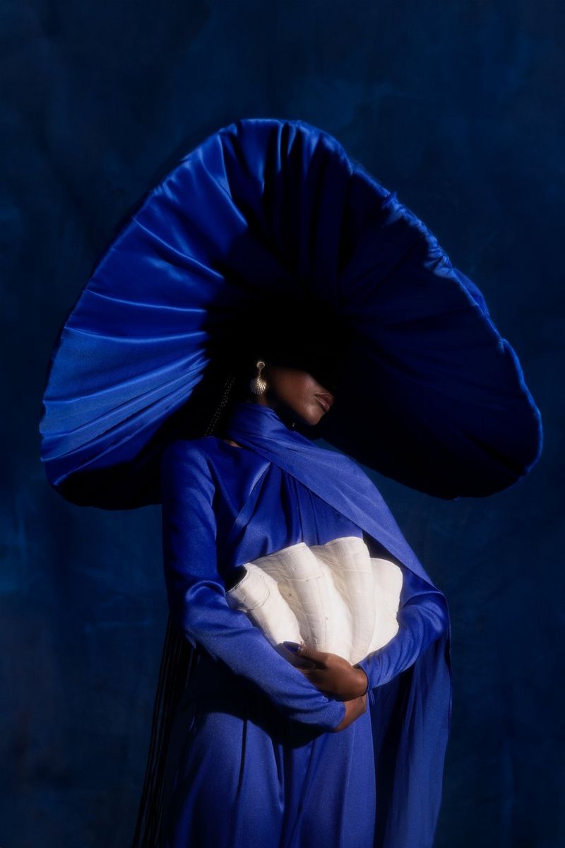 A model, wearing a bright blue satin dress and a huge wide-brimmed hat, with her arms around a large shell. Taken by Nina Zimolong on a Canon EOS R5.