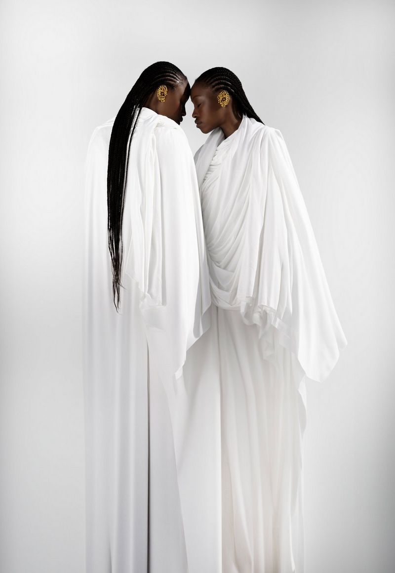 Two women in long, white flowing robes stand facing each other with their foreheads touching, in a portrait taken by Nina Zimolong on a Canon EOS R5.