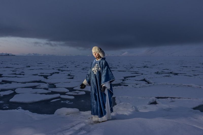 A female pastor, in long blue robes and a fur hat, standing on a floating field of sea ice.