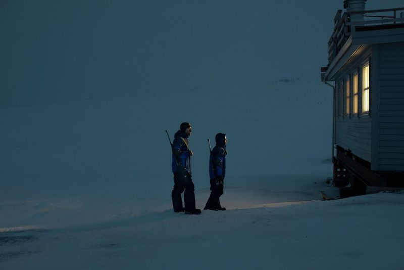 Deux silhouettes vêtues de vêtements chauds et portant des fusils parcourent un paysage sombre et enneigé en direction d'un bâtiment.