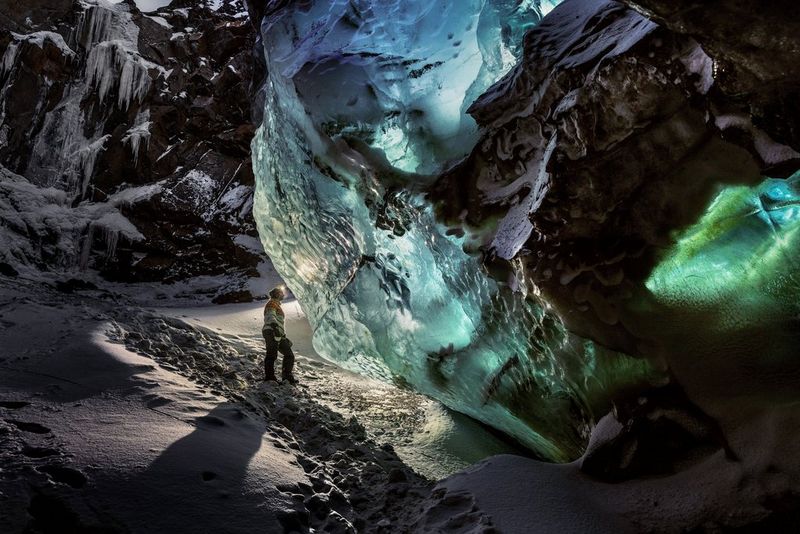 A hiker standing in an ice cave beside a huge wall of ice lit green and blue.