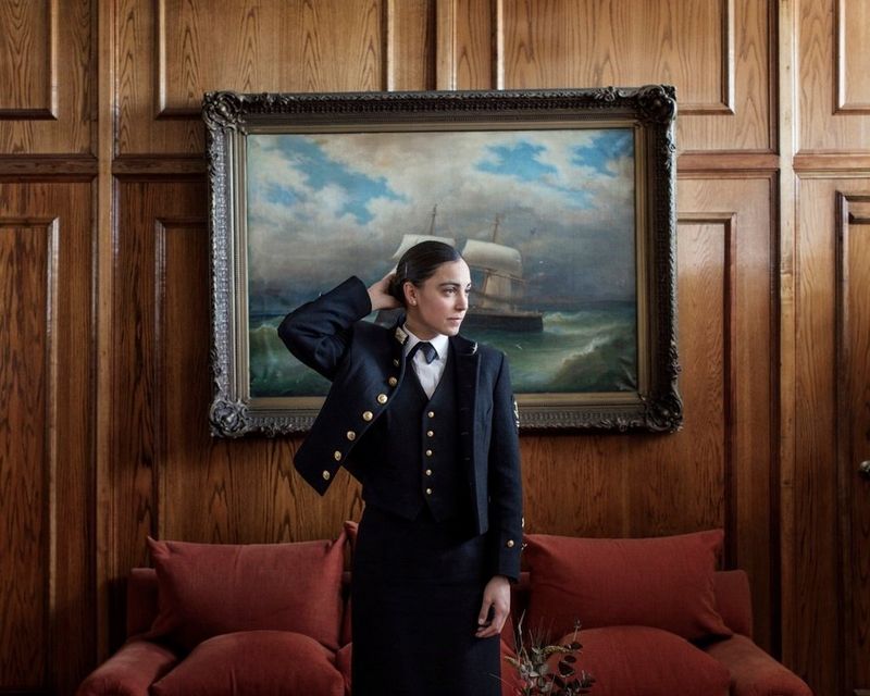 Une jeune femme en uniforme devant une peinture nautique, sa main au dos de ses cheveux.