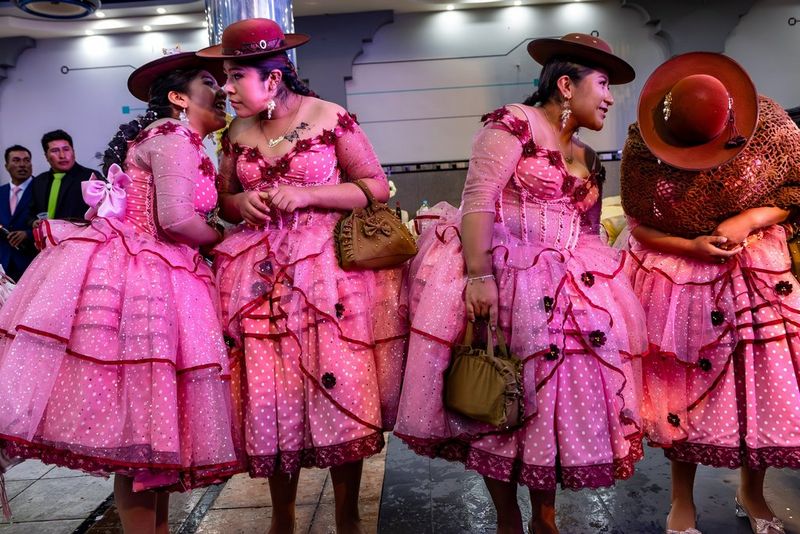 Four Aymara women, in matching pink polka dot dresses and red hats, talking amongst themselves as they stand in a line.