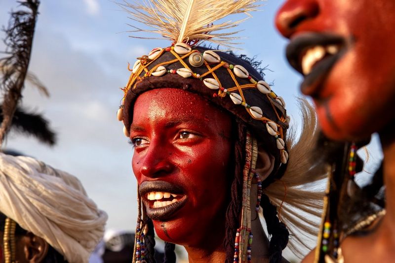 A young man wearing red face paint and a head adornment decorated with shells and a large white feather. 