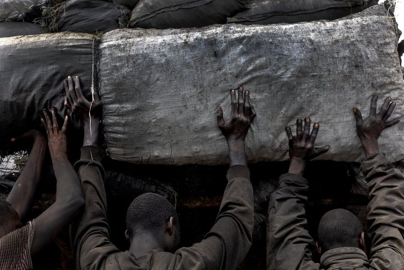 A row of men, with their backs to the camera and just their heads and arms visible, loading bags of charcoal onto the back of a truck.
