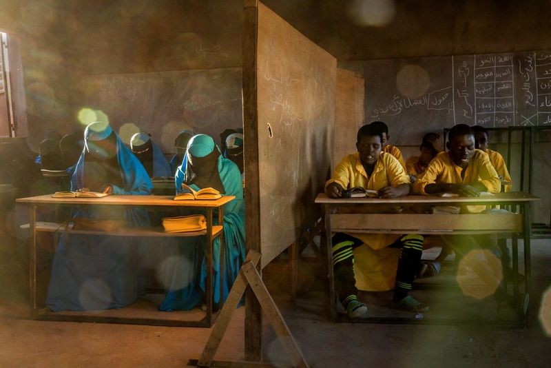 Male and female students in colourful robes sit behind desks in a classroom that's divided in half.