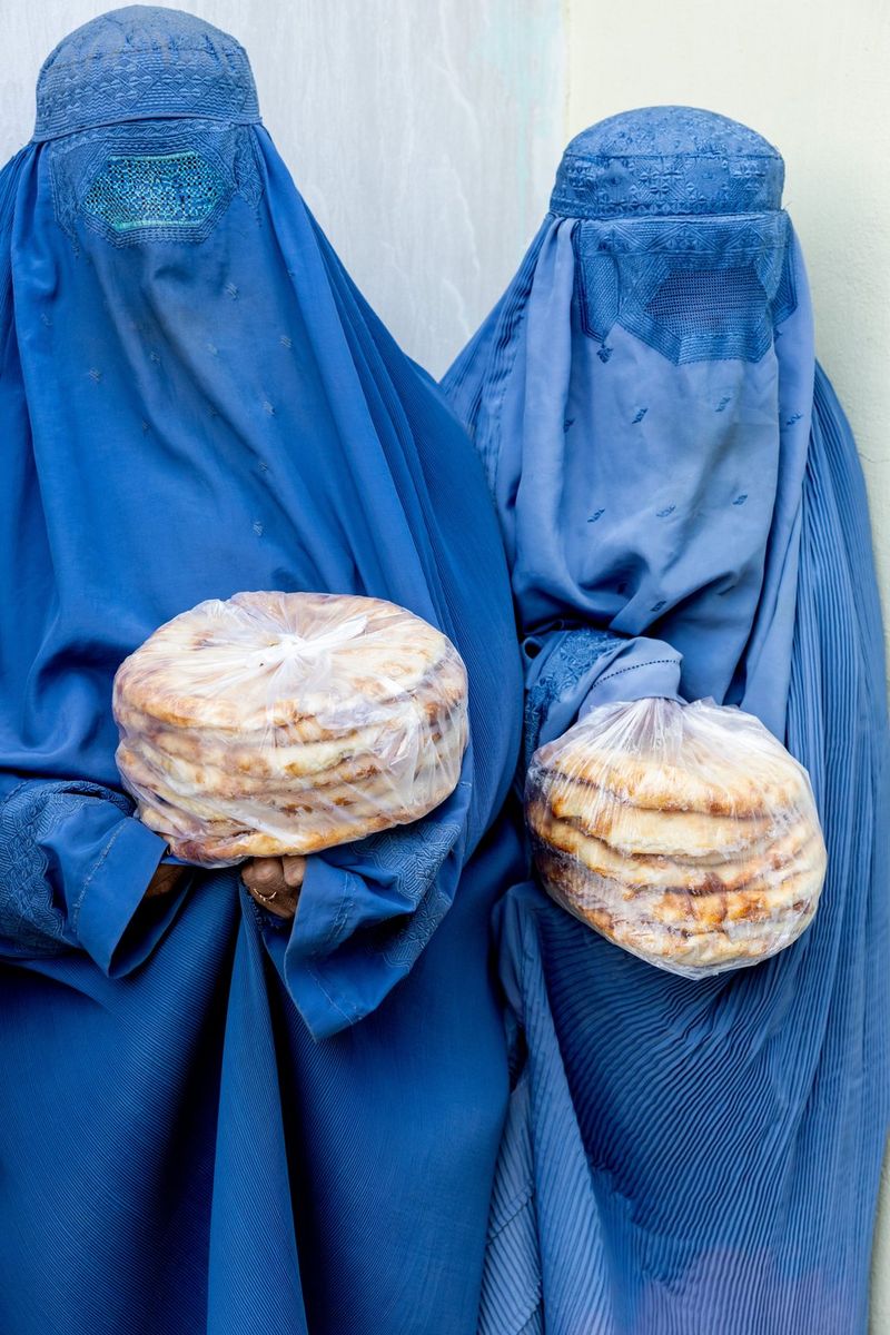 Two women in bright blue burkas holding plastic bags filled with flatbreads.