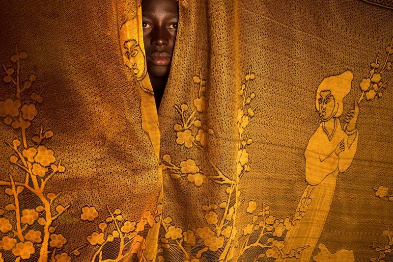 A young Dogon woman peers out from between two bright yellow patterned curtains.