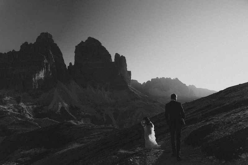 A black and white photograph of a bride and groom, shot from behind, walking across a rocky landscape.