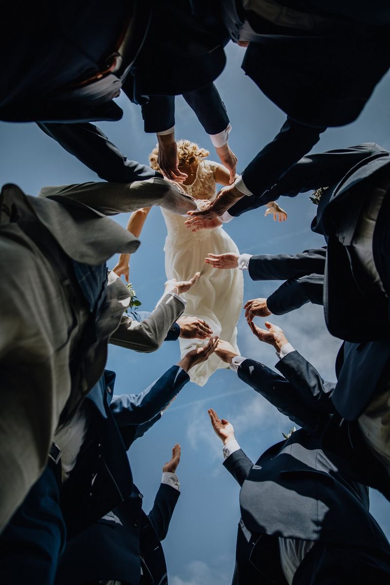 Shot from below, a bride in a long, white dress is thrown into the air by a circle of groomsmen. Taken on a Canon EOS 5D Mark IV by Raïs De Weirdt.