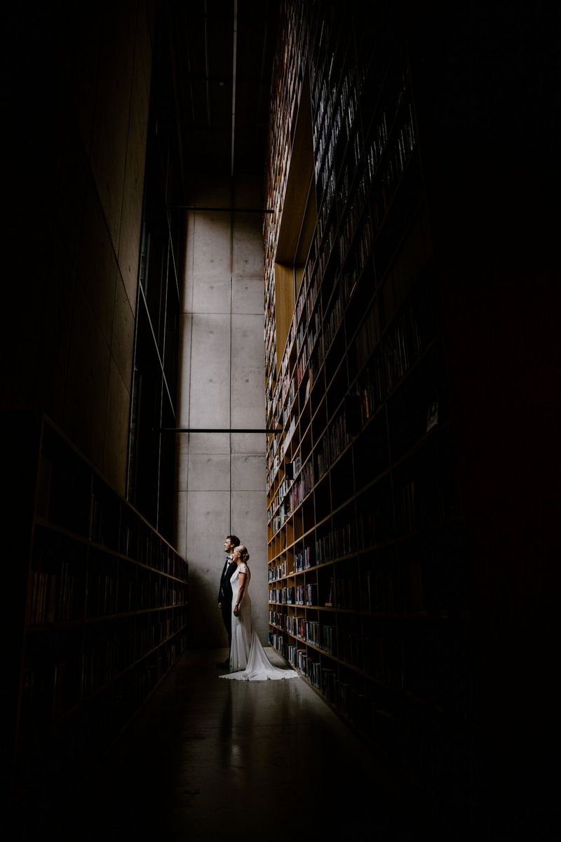A newlywed couple stand facing a large window, their backs towards floor-to-ceiling shelves of library books. Taken on a Canon EOS 5D Mark IV by Raïs De Weirdt.