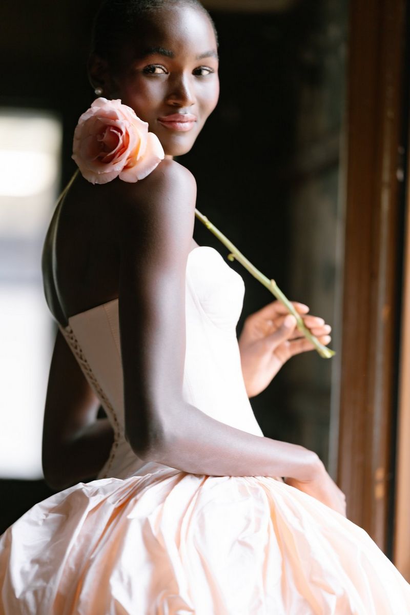 A portrait of a woman posing in a peach strapless bridal dress and holding a peach rose up to her face.