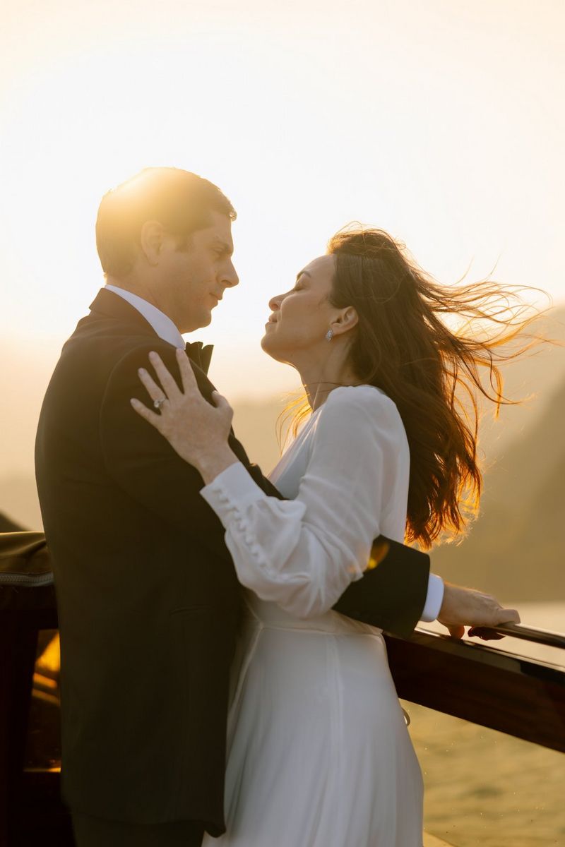 A bride in a long-sleeved white dress embracing her groom in front of a low sun.