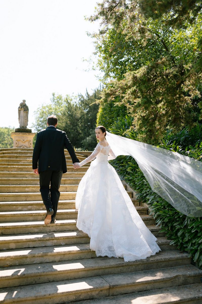 A bride in a long white dress and veil looking back as she walks with her groom up a flight of stone steps.