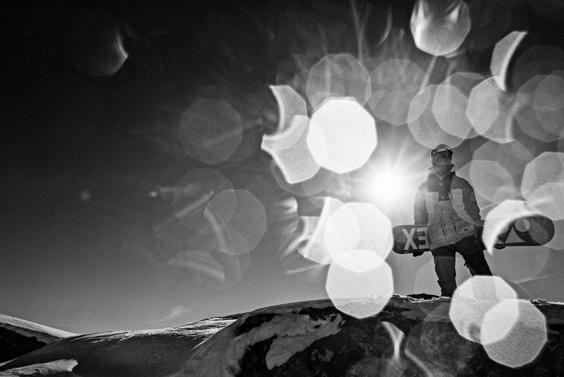 Snowboarder Tim-Kevin Ravnjak stands at the top of a Swiss mountain, surrounded by lens flare from the sun.