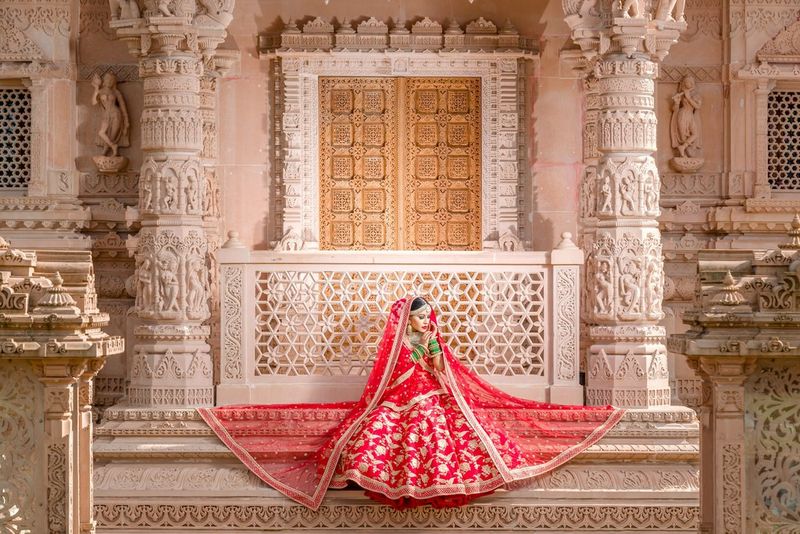 A bride in a red and gold dress sits on the steps of an ornate Hindu temple, with a long veil spread out around her.