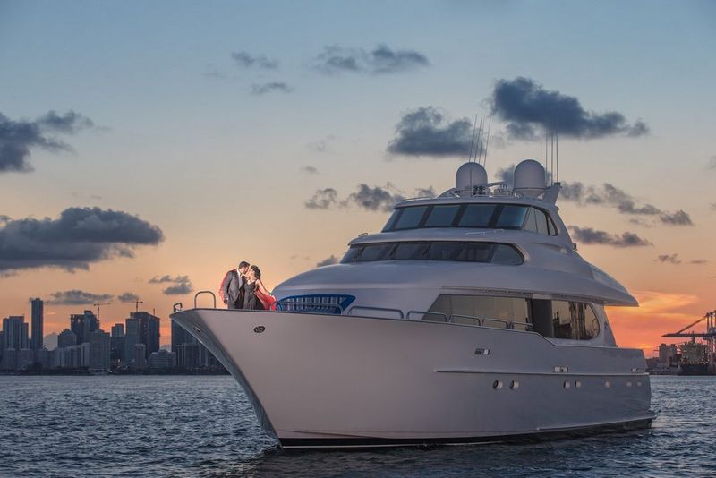 A bride and groom embrace at the front of a luxurious yacht as the sun sets behind them.