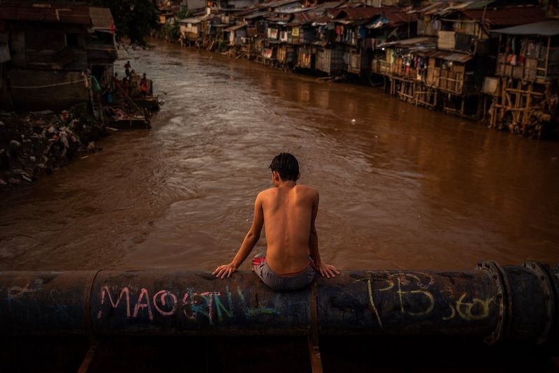 A young man sitting on a metal pipe above the muddy waters of the Ciliwung River in Jakarta, Indonesia. Taken on a Canon EOS 5D Mark IV by Canon Ambassador Santi Palacios.