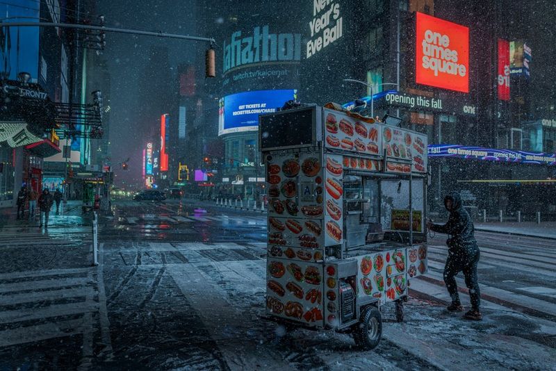 A man pushing a food cart along a street in New York as snow falls at first light.