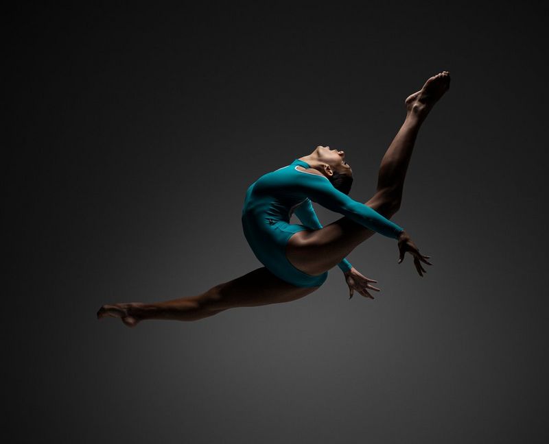 A ballet dancer in a turquoise leotard leaping into the air with her arms thrown back and her legs outstretched.
