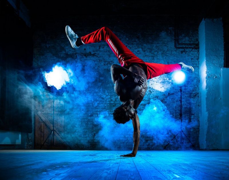 A shirtless breakdancer in red trousers balances athletically on one hand in a blue-lit, smoky room.
