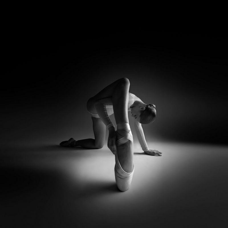 A black and white portrait of a ballet dancer kneeling down with one foot forward en pointe.