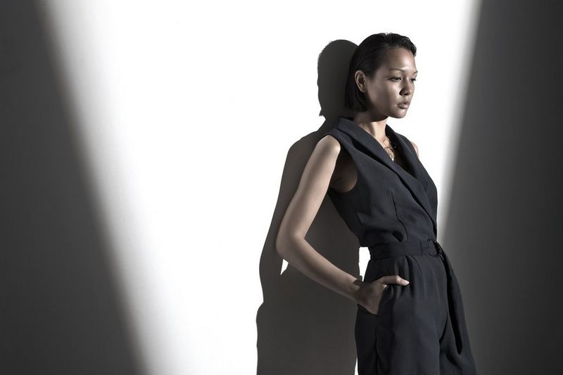 A female model wearing a black jumpsuit stands with her hands in her pockets against a carefully lit wall. 