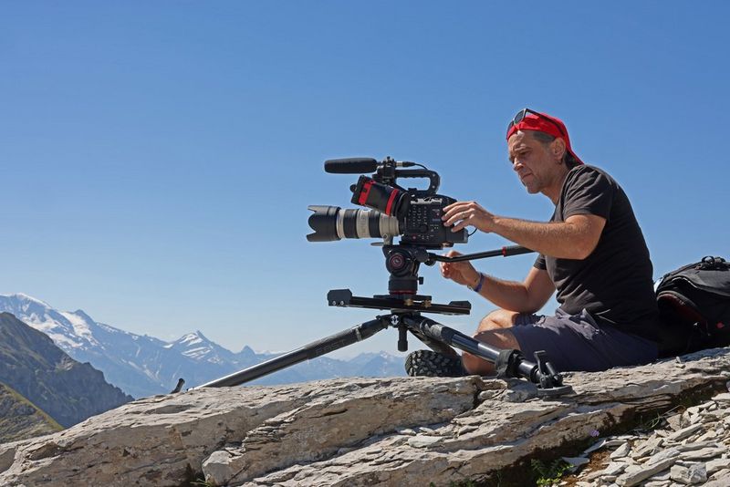 Canon Ambassador Sébastien Devaud adjusts his Canon video camera while sitting on a rocky outcrop beneath a bright blue sky.