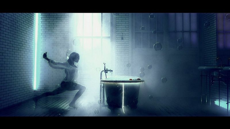 A dancer lunges towards an overflowing bath, one leg behind her and with her arms outstretched, in a smoke-filled industrial-style setting. Bubbles appear to be floating in the air around her.