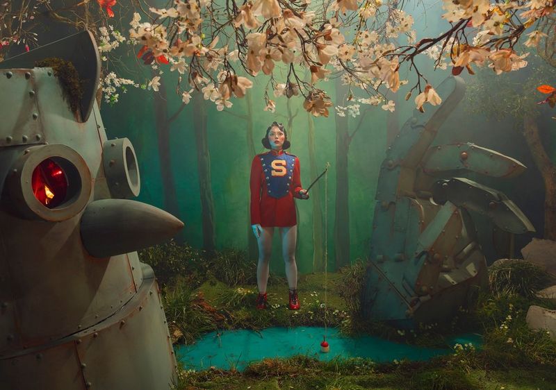 A stylised portrait of a woman in a red tunic holding a fishing rod, framed by trees, both real and fake, and with a robot in the foreground. Taken on a Canon EOS 5DS by Canon Ambassador Sébastien Salamand dit Le Turk.