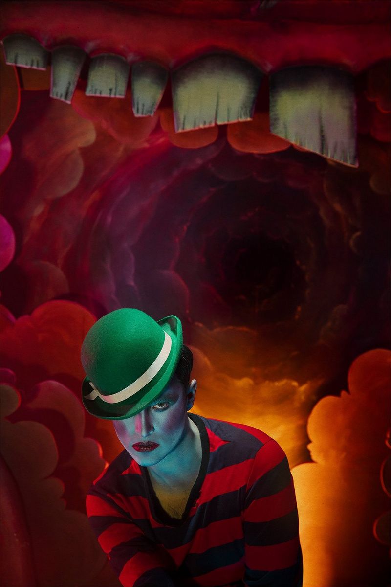 A heavily made-up model wearing a striped top and a green bowler hat poses in front of a red, cave-like backdrop. Taken on a Canon EOS 5DS by Canon Ambassador Sébastien Salamand dit Le Turk. 