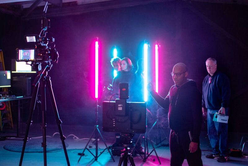 A film set lit by neon pink and blue lights. Two actors in the centre and two film crew to their right. 