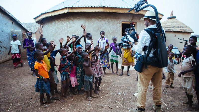 A man filming a group of waving children with a Canon EOS Cinema camera.