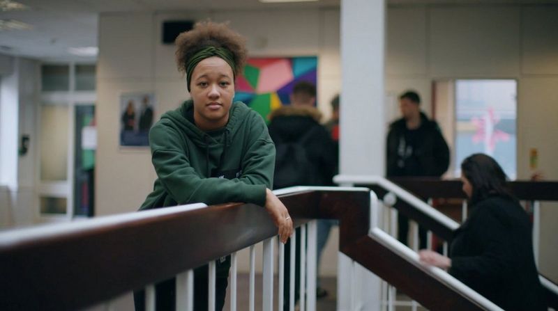 A young woman leans on a bannister, looking directly at the camera. Other young people can be seen in the background.