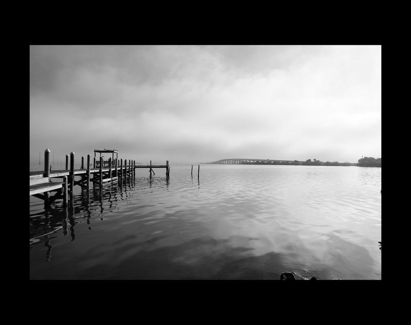 Une image en noir et blanc d'un plan d'eau immobile par temps nuageux. Une petite jetée couverte d'oiseaux est présente à gauche de l'image.