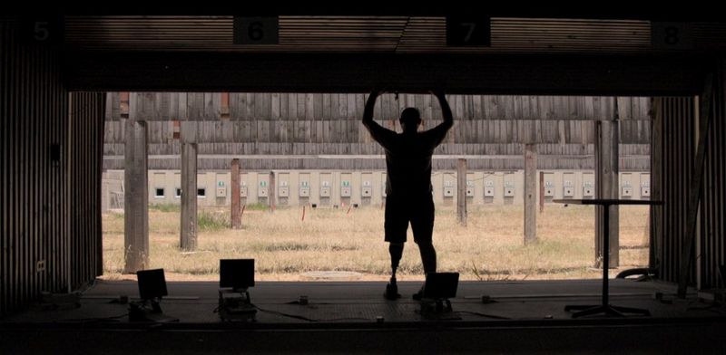 A still from cinematographer Tanya Aizikovich's showreel, showing a man with one false leg holding on to an opening garage-style door, staring out at rows of wooden supports. © Tanya Aizikovich