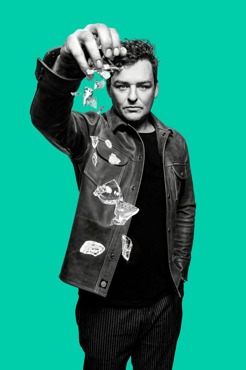  A portrait of Czech artist Martin Janecký against a plain green background, shards of glass falling from his raised hand. Taken on a Canon EOS 5DS by Tomáš Třeštík.