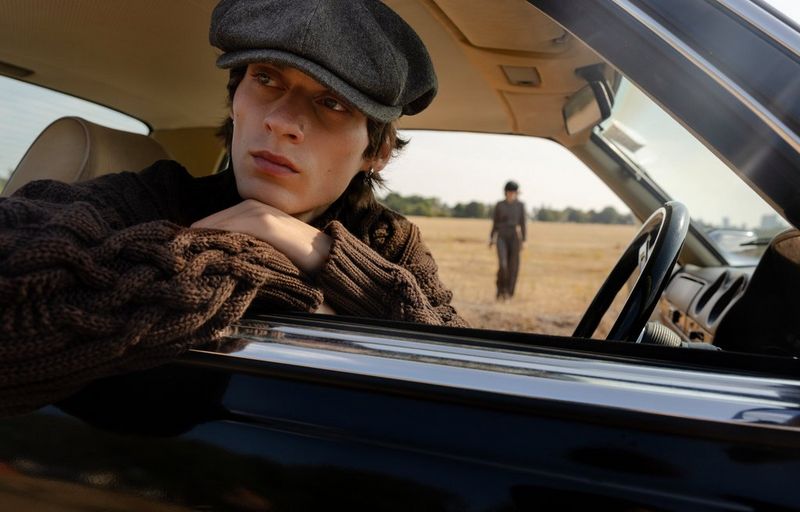 A fashion portrait of a male model in a flat cap and woollen jumper leaning on the windowsill of the car he is sitting at the wheel of.