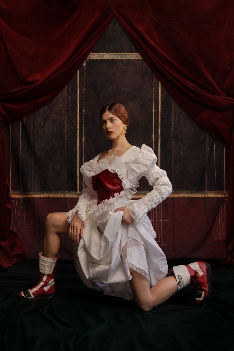A woman in a white ruffled dress and red leather boots posing on one knee in front of a dark, panelled backdrop.